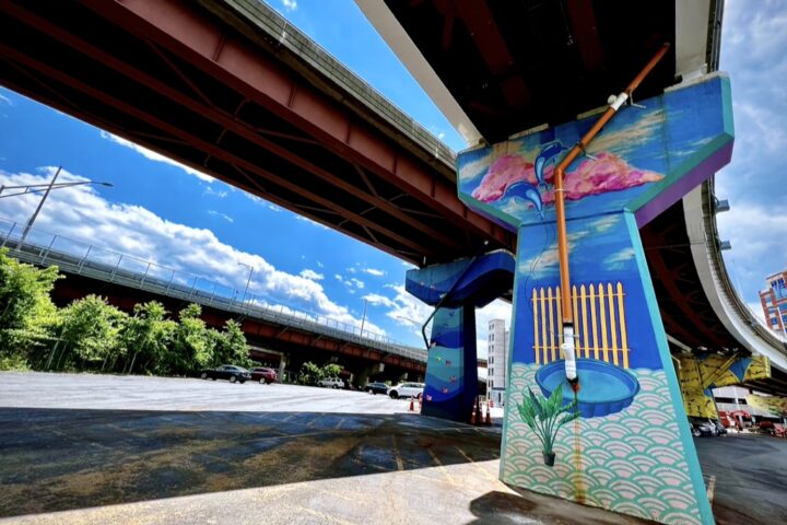 Colorful mural painted on a highway support pillar under an elevated roadway in a parking lot.
